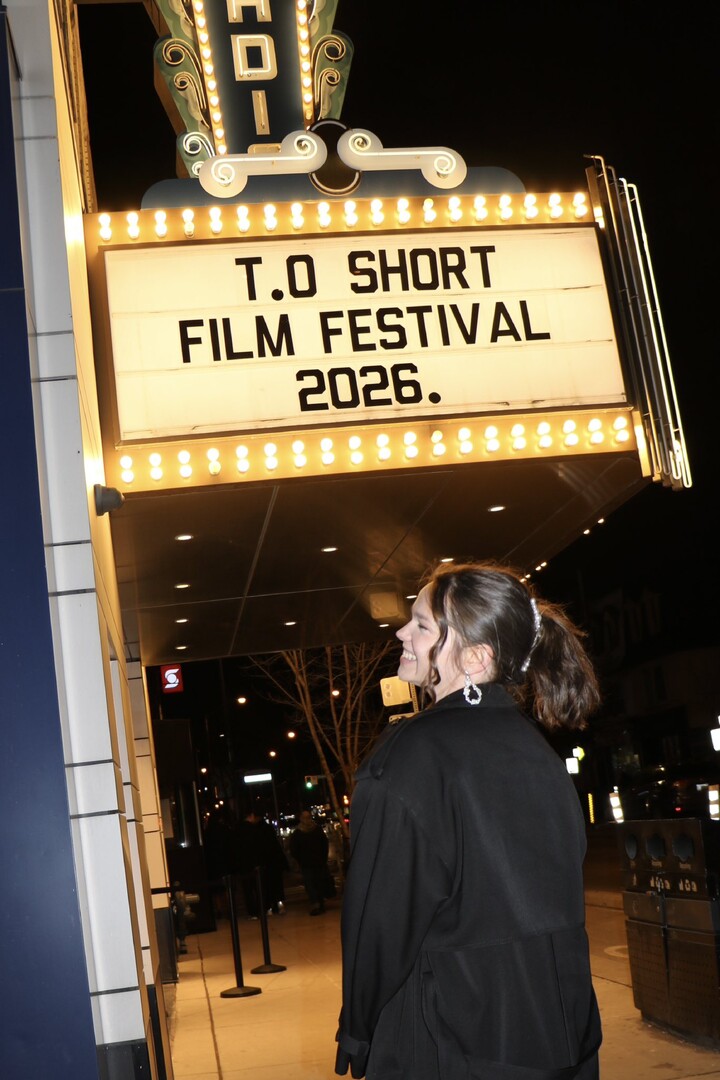 A filmmaker smiles up at the glowing T.O Short Film Festival 2026 marquee on a city sidewalk at night.