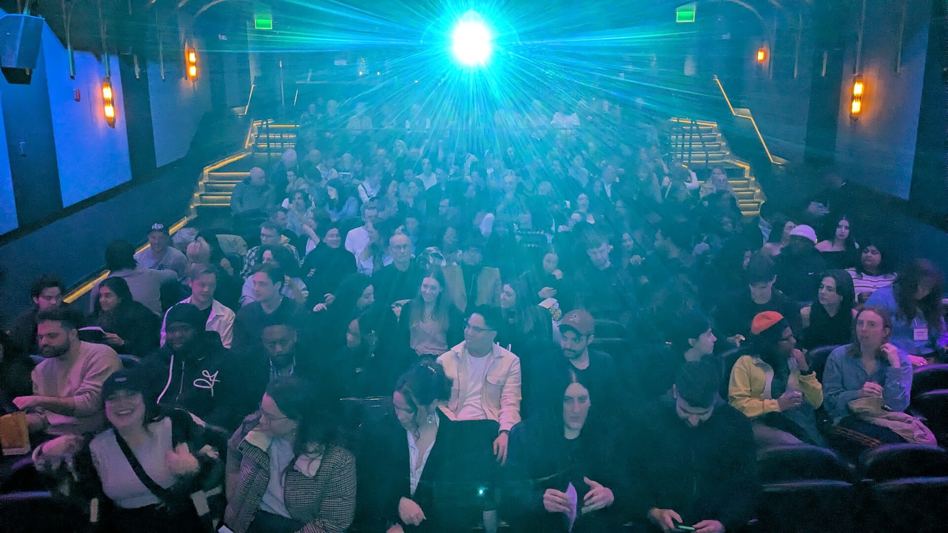 Projector light flares across a full cinema auditorium, seen from the screen towards the back of the house.