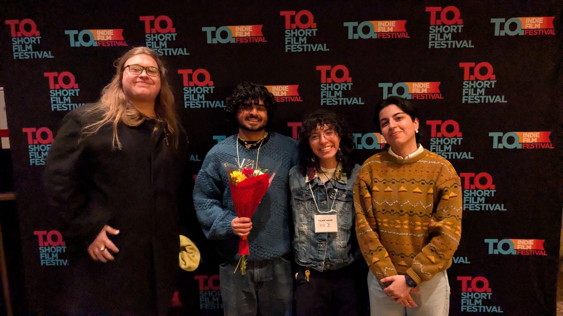 Four filmmakers pose in front of a T.O Short Film Festival backdrop; one holds a wrapped bouquet.