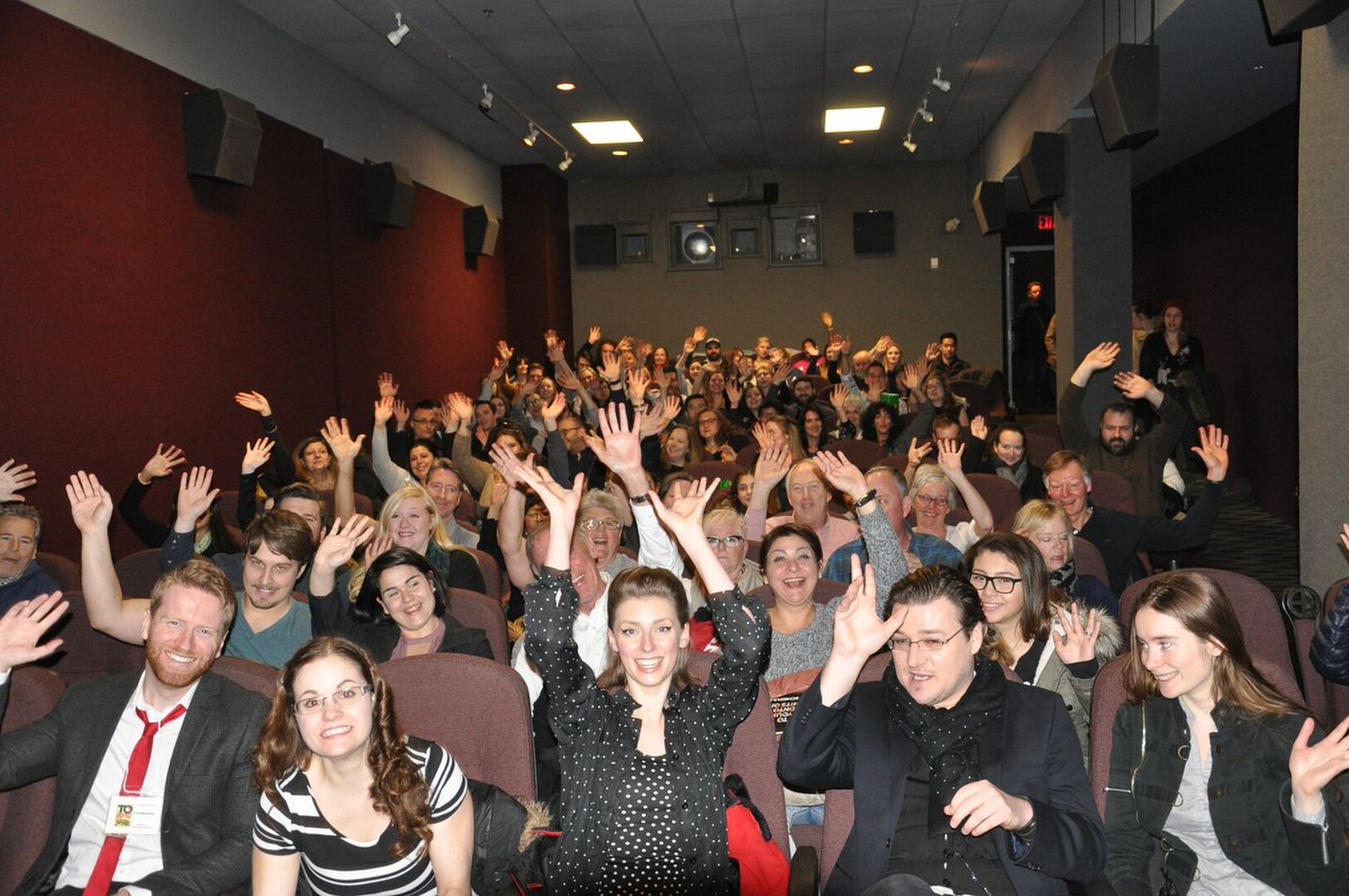 A seated audience in maroon cinema seats faces the camera with hands raised and smiles.