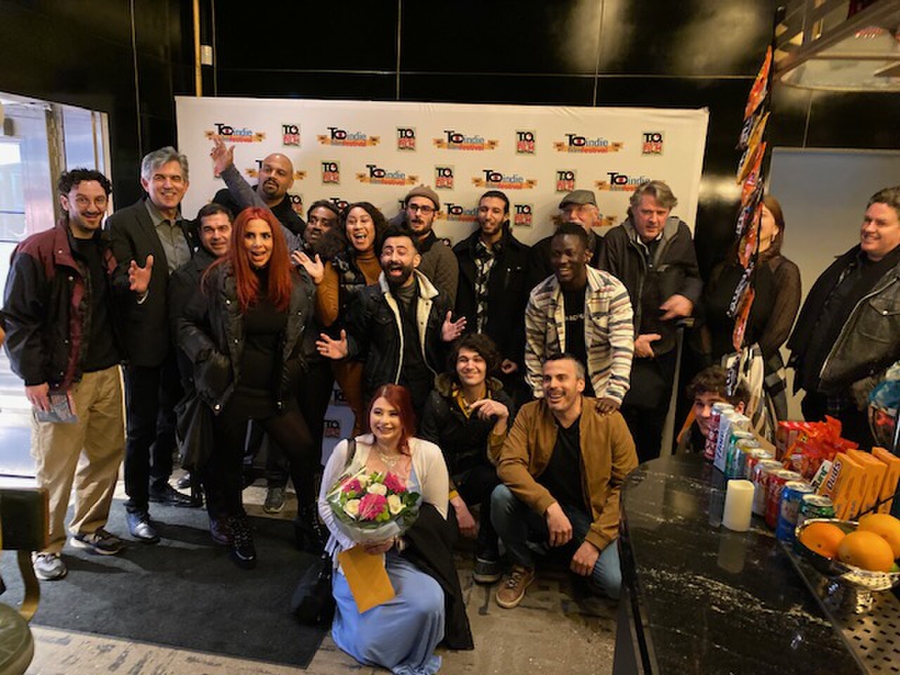 A large group of about twenty people pose in front of a white T.O Indie Film Festival backdrop, a woman kneeling in front holding a bouquet.