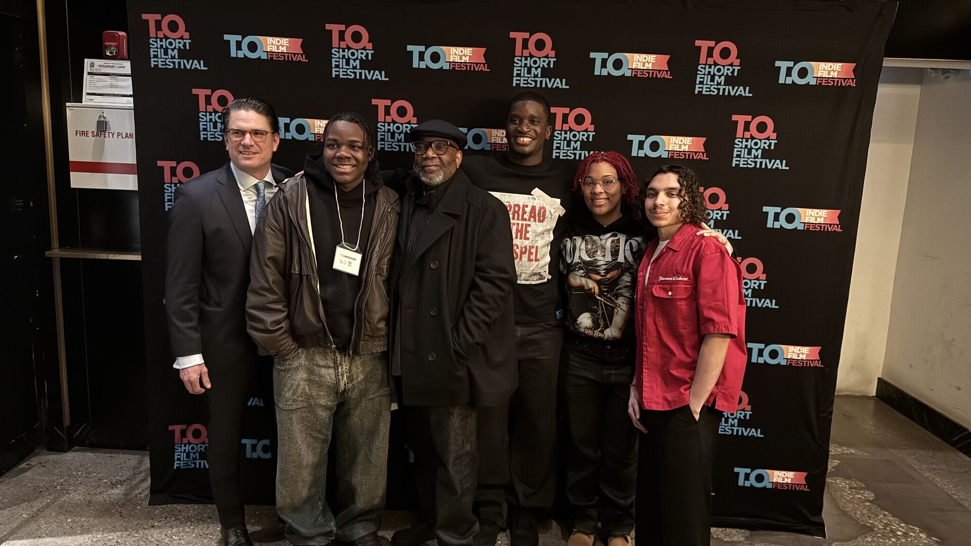 Six filmmakers stand together in front of a black T.O Short Film Festival backdrop; one wears a festival Filmmaker badge.