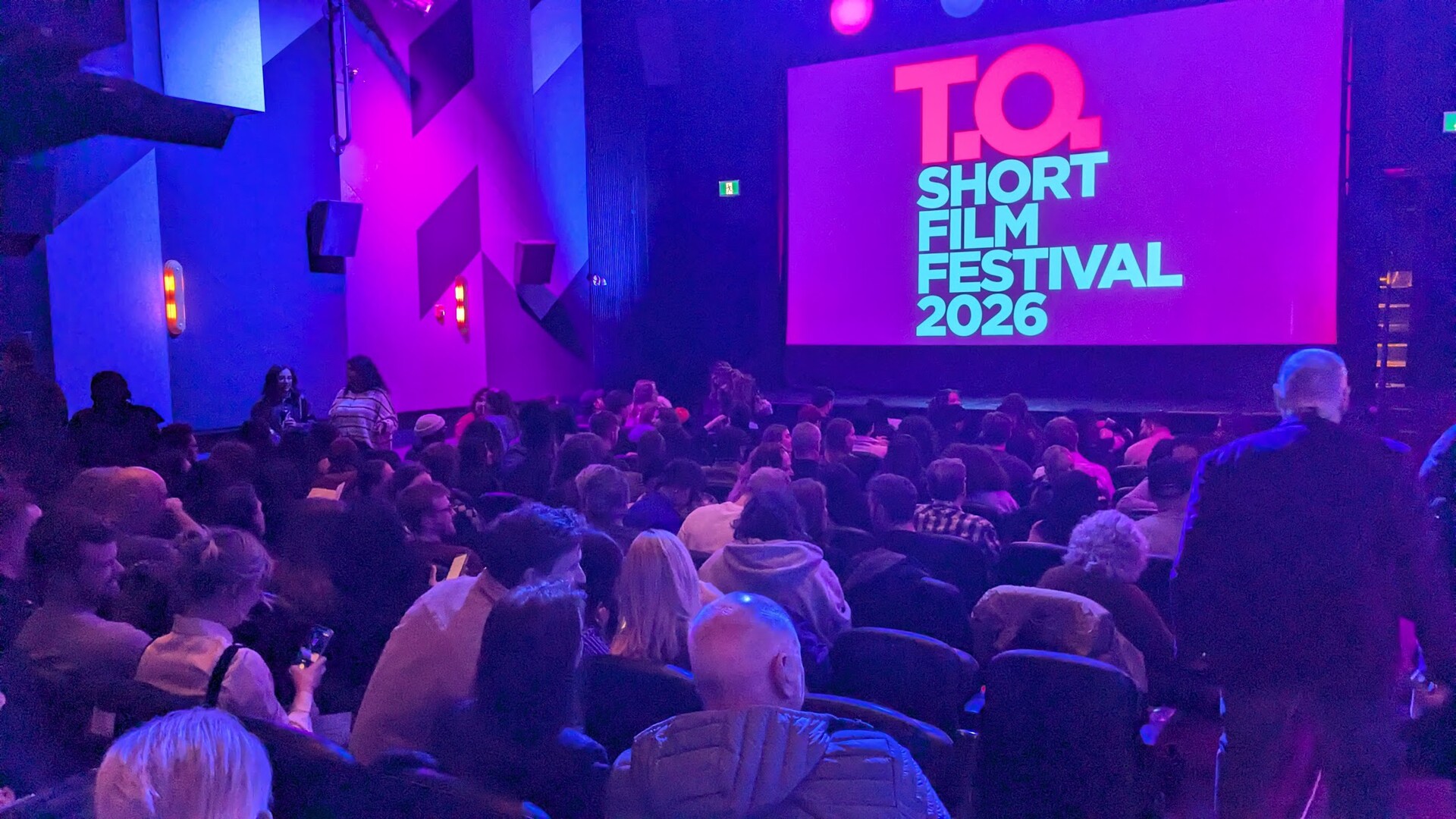 A packed auditorium seen from behind the seats, facing the illuminated festival screen under pink and blue light.